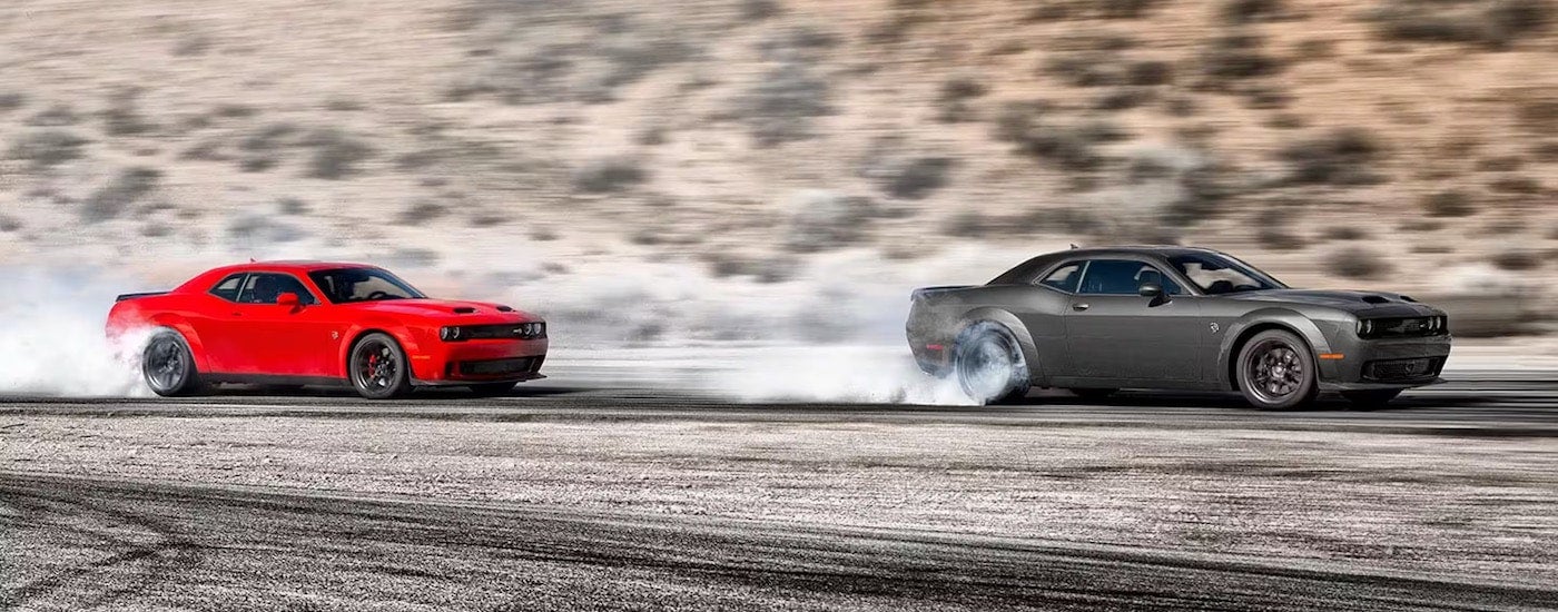 A red and a grey 2023 Dodge Challenger Hellcat Widebody are shown from the side while doing burnouts after leaving a Dodge dealer.